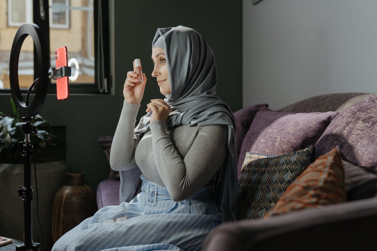 A woman in hijab applying makeup indoors, recording a tutorial with a smartphone and ring light.